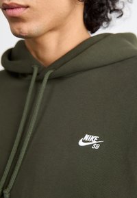 Olive green hoodie with a drawstring. Features a small white Nike SB logo on the chest. Fabric appears soft and textured.