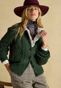 Sötét zöld, texturált mintázatú kötött kardigán, fehér fodros blúzzal és kockás nadrággal párosítva. Kiegészítve bordó kalappal.