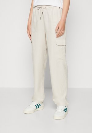 Personne portant un pantalon cargo beige ample avec une taille à cordon et des baskets blanches avec des rayures vertes, se tenant contre un fond uni.