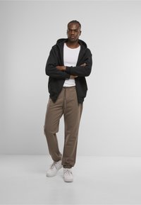 Schwarze Zip-Up-Hoodie über einem weißen T-Shirt, kombiniert mit beigen Sweatpants und weißen Sneakers. Glattes Material mit lockerer Passform.