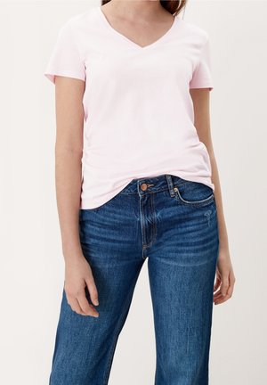 Femme portant un t-shirt rose clair à manches courtes et col en V et un jean bleu en denim, debout devant un fond clair uni avec une posture détendue.