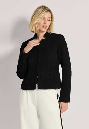 Blonde Frau trägt eine strukturierte schwarze Jacke und weiße Hose mit schwarzen Seitenstreifen und steht vor einem schlichten Hintergrund.