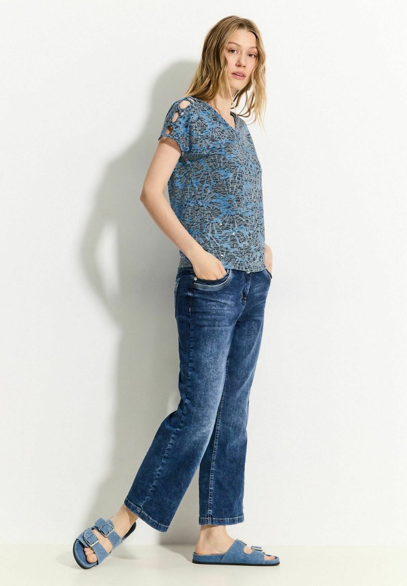 Maglietta a maniche corte blu con motivo e scollatura a V, abbinata a jeans in denim blu sbiaditi e sandali blu con fibbie regolabili.
