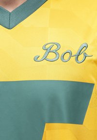 Κίτρινη και πράσινη αθλητική φανέλα με τιρκουάζ γιακά και κεντημένο όνομα «Μπομπ» στο στήθος.