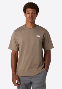 Mies, joka on pukeutunut taupe-väriseen, lyhythihaiseen T-paitaan, jossa on The North Face -logo rinnassa, ja harmaisiin housuihin, seisoo käsi taskussa.