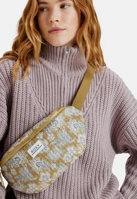 Beige vyöbag, jossa on kukkakuosi ja kultainen vetskari, kullakin rokkaan kropan yli teksturoidun laventelivärisen neuleen päällä, jossa on korkea kaulus.