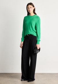 Πράσινος πλεκτός πουλόβερ με μακριά μανίκια, συνδυασμένος με φαρδύ μαύρο παντελόνι. Κρατάει μια μαύρη τσάντα χεριού, που έχει απλές γραμμές και δεν διαθέτει σχέδια.