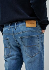 Jasnoniebieskie dżinsy z denimu z brązową skórzaną naszywką oznaczoną "Salsa Jeans". Posiadają podwójnie szyte tylne kieszenie oraz gładkie, teksturowane wykończenie.