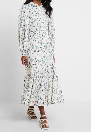 Femme portant une longue robe blanche à motifs floraux avec des manches longues et des sandales blanches à brides, debout devant un fond uni.