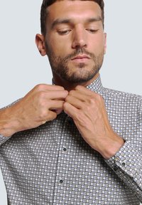 Overhemd met knoopsluiting en een geometrisch patroon in blauwtinten en beige, lange mouwen en een kraag. Model is de kraag aan het aanpassen.