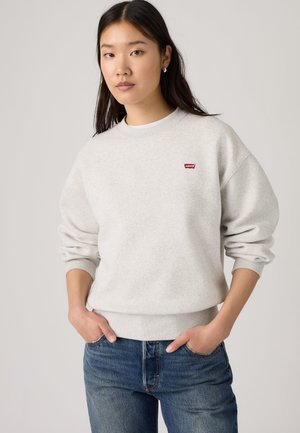 Junge Frau trägt einen hellgrauen Levi's-Sweatshirt und blaue Jeans mit den Händen in den Taschen, stehend vor einem schlichten hellen Hintergrund.