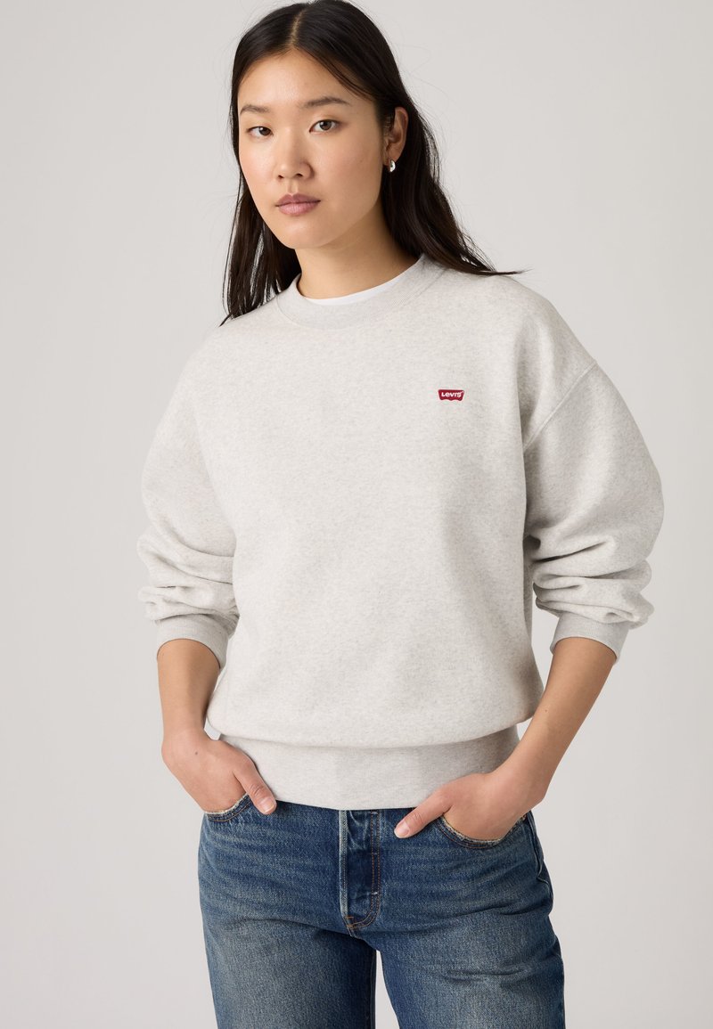 Νέα γυναίκα φοράει ανοιχτό γκρι φούτερ Levi's και μπλε τζιν με τα χέρια στις τσέπες, όρθια μπροστά σε ένα απλό φωτεινό φόντο.