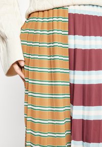 Taille et main d'une personne portant une jupe plissée à rayures horizontales moutarde et marron, associée à un pull en maille blanc.