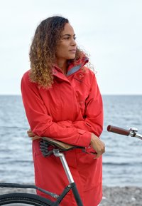 Veste imperméable rouge avec des accents gris, se tenant à côté d'un vélo avec une selle et des poignées en cuir marron, sur fond de mer.