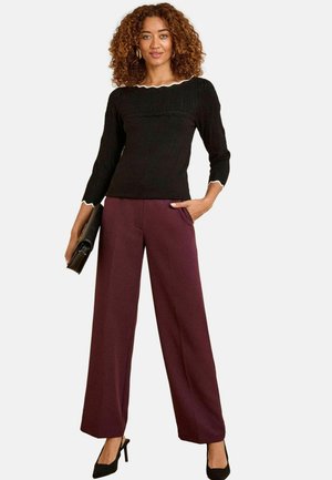 Eine Frau mit lockigem Haar trägt einen schwarzen Pullover und bordeauxfarbene weite Hosen, hält eine schwarze Clutch und steht selbstbewusst.