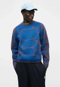 Man wearing a blue patterned sweater, light blue cap, and black Nike pants, looking to the side with hands in pockets against a plain background.