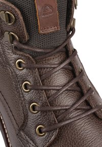 Bota de cuero marrón con acabado texturizado, cordones marrones, ojetes de metal y un puño de tela. Se adjunta una pequeña etiqueta de cuero cerca de la parte superior.