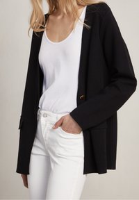 Black cardigan with a relaxed fit, featuring two front pockets and a single gold button, paired with a white tank top and white jeans.