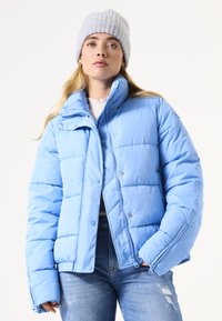 Lichtblauwe pufferjack met een ritssluiting en drukknoopjes, met een hoge kraag en gestikt textuur. Gecombineerd met versleten jeans.