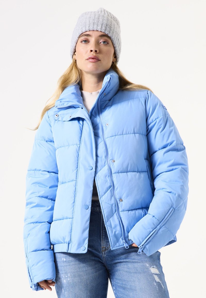 Lichtblauwe pufferjack met een ritssluiting en drukknoopjes, met een hoge kraag en gestikt textuur. Gecombineerd met versleten jeans.