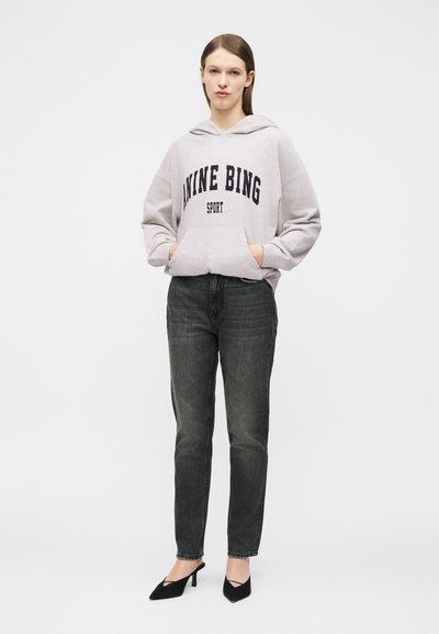 Woman standing with hands in pockets wearing a light grey hoodie with "NINE BING SPORT" text, black jeans, and black pointed heels.