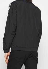 Svart bomberjacka med en slät, matt yta; har vita sidostråk och elastiska muddar och nederkant. Bärs över svarta byxor.