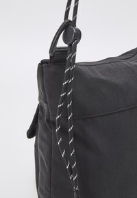 Černý látkový sáček v rohu s nastavitelným proužkovaným stahovacím šňůrkem a černým plastovým sponou na světlem pozadí.