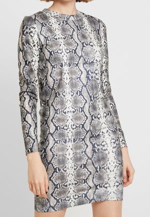 Žena v dlouhých rukávech a krátkých šatech se vzorem šedé a černé hadí kůže, stojící před jednoduchým světlým pozadím.