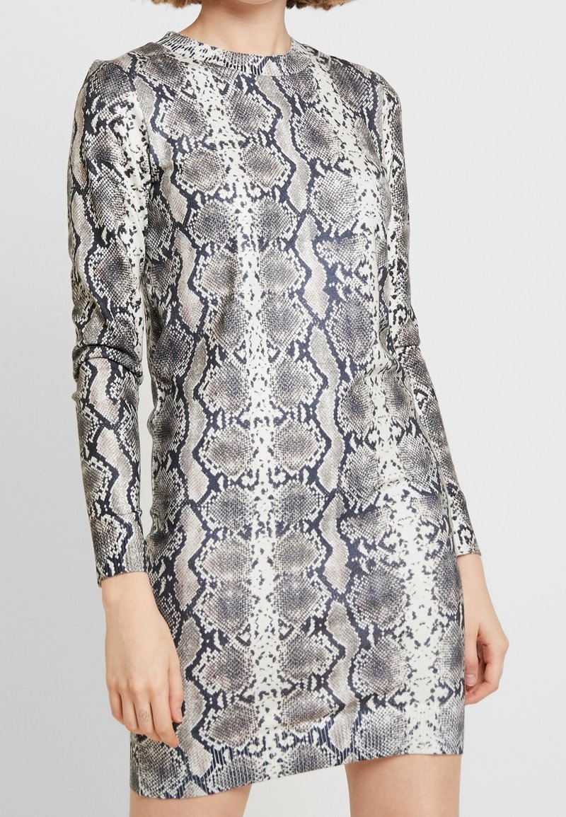 Femme portant une robe courte à manches longues avec un motif peau de serpent gris et noir, debout devant un fond clair uni.