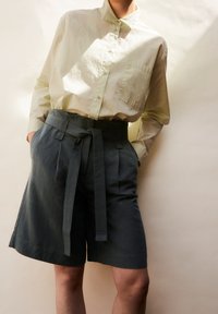 Světle zelená košile s límečkem, knoflíky a náprsní kapsou, spojená s tmavě šedými šortkami s vysokým pasem, které mají uvázaný pásek a sklady.