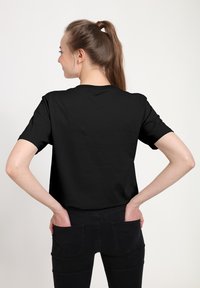 Svart kortärmad t-shirt med rund halsringning, gjord av ett slätt tyg. Modellen står med ryggen mot kameran, med händerna i fickorna, och visar upp ryggdesignen.