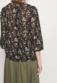 Svart transparent blus med blommönster i gult, vitt och rött. Har trekvartslånga ärmar och en rund halsringning, bärs över en grön kjol.