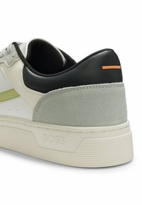 Gros plan sur le talon d'une basket avec une semelle blanche, du daim gris, un col en cuir noir, une bande vert clair et un accent orange à l'arrière.