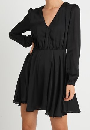 Femme portant une robe noire à manches longues, col en V, taille cintrée et jupe évasée au-dessus du genou.