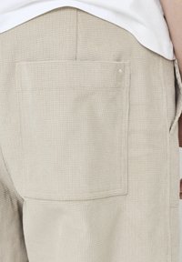 Pantalons texturés beiges avec un motif à carreaux en relief, une poche arrière visible et un ourlet soigné. Fabriqués en tissu léger.