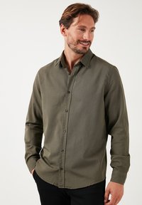 Camisa de botones verde oliva de manga larga, hecha de un tejido texturizado. Presenta un cuello clásico y botones negros en la parte delantera.