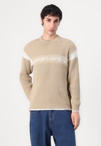 Beige Ripppullover mit weißem, horizontalem Streifen und Logo auf der Brust. Er hat lange Ärmel, einen runden Halsausschnitt und einen lässigen Schnitt.