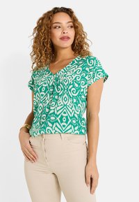 Blusa verde con estampado, escote en V y mangas cortas, con diseños abstractos en blanco; combinada con pantalones ajustados de un beige claro.