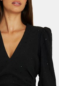 Schwarzes Kleid mit tiefem V-Ausschnitt, strukturiertem Stoff, verziert mit kleinen schwarzen und silbernen Pailletten sowie Puffärmeln.