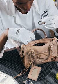 Personne portant des gants blancs et une blouse de laboratoire inspecte un sac à main en cuir beige à l'aide d'une loupe sur un tapis de découpe.