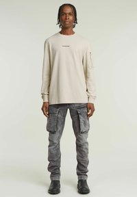 Beżowa koszula z długim rękawem o diamentowej strukturze i logo; noszona z szarymi spodniami cargo z wieloma kieszeniami i czarnymi butami.