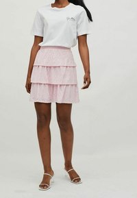 Jupe rose à couches avec motif ajouré, ceinture élastique, associée à un shirt à manches courtes blanc avec un imprimé noir. Sandales blanches à talons.