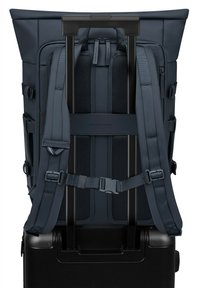 Sac à dos marine avec des bretelles réglables et un rembourrage en maille, conçu pour s'adapter sur une valise noire à roulettes. Tissu lisse avec des accents minimaux.