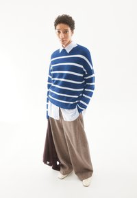 Blau-weiß gestreifter Pullover über einem weißen Hemd mit Kragen, kombiniert mit beigen weiten Hosen. Das Model hält einen kastanienbraunen Artikel. Cremefarbene Schuhe.