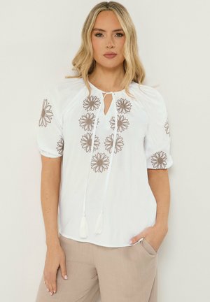 Femme blonde portant un chemisier blanc avec une broderie florale marron et un pantalon beige, debout avec une main dans la poche.