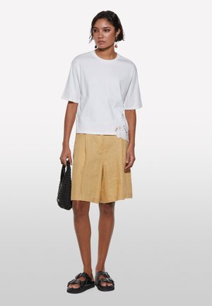 T-shirt en coton blanc à manches courtes, avec un détail en dentelle à l'ourlet. Associé à un short en lin jaune clair et des sandales noires.