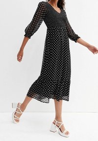 Schwarz gepunktetes Midi-Kleid mit durchsichtigen langen Ärmeln, V-Ausschnitt und gestuftem Rüschensaum. Kombiniert mit weißen Strappy-Blockabsatzsandalen.