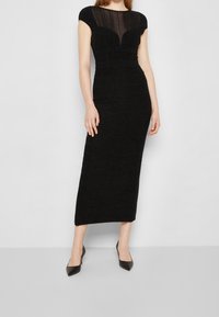Robe midi noire avec des manches courtes, présentant un panneau supérieur transparent, un tissu texturé et une taille cintrée. Portée avec des escarpins noirs à bout pointu.