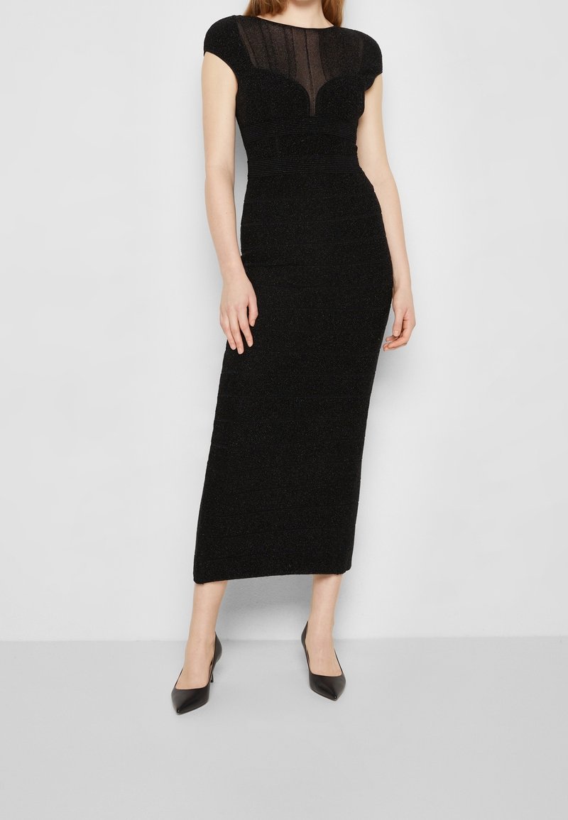 Robe midi noire avec des manches courtes, présentant un panneau supérieur transparent, un tissu texturé et une taille cintrée. Portée avec des escarpins noirs à bout pointu.