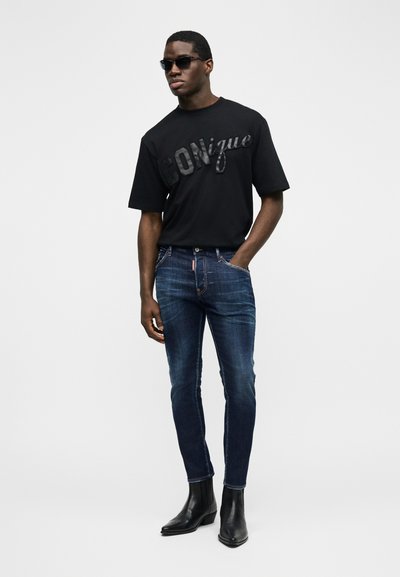 Homme portant des lunettes de soleil noires, un t-shirt noir "ICONique", un jean bleu foncé et des bottines noires, debout avec une main dans la poche.